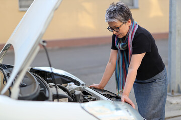 A frustrated mature female driver stands beside her broken-down car, seeking assistance for engine trouble on the roadside.