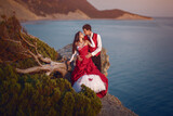 Fototapeta  - Wedding photo shoot of a beautiful bride and groom in red dresses on a mountain overlooking the ocean, happy holiday of the newlyweds.