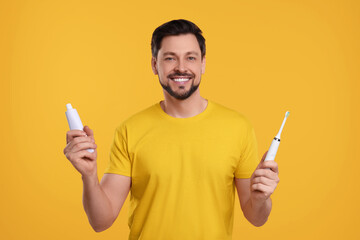 Wall Mural - Happy man holding electric toothbrush and tube of toothpaste on yellow background