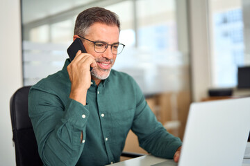 middle aged businessman making call on cellphone using laptop in office. smiling busy mature old pro