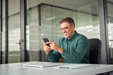 Smiling mid aged businessman executive using cell phone at work desk. Happy busy mature older professional business man manager investor checking finance apps on smartphone in office looking at mobile