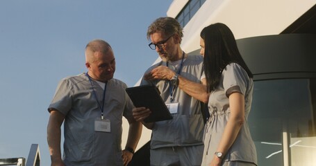 Medical staff in uniform stand outdoors at hospital entrance. Mature doctor talks with colleagues, shows medical tests results using digital tablet. Modern clinic or medical center. Healthcare system.