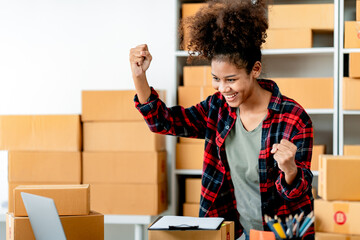 Wall Mural - Young African woman prepares parcel boxes and checks online orders for delivery to customers on laptop. Shipping and online shopping concepts