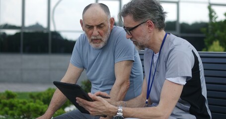 Wall Mural - Mature doctor with tablet in hands sits with elderly patient on the bench and consults him. Professional medic in uniform makes diagnosis to client using digital tablet. Medical staff work outdoor.