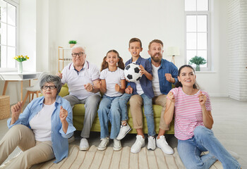 Happy big family watching soccer match on TV in living room at home. Cheerful grandparents, mother, father and two their cute children football fans cheering up support favorite team with soccer ball