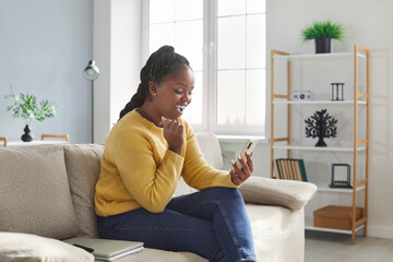 Excited young cheerful african happy girl using mobile phone while sitting on sofa at home. Woman resting on couch with cellphone and looking at screen, chatting or reading pleasant sms message.