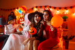 Portrait of two cheerful women in carnival costumes having fun during Halloween celebration at home. Women dressed as witch and devil sitting on sofa with balloon and pumpkin on background of people.