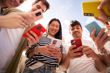 Low angle view multiracial group young generation z in circle using phones together outdoors. Cheerful student friends looking at mobile enjoying social media content. People and technology addiction 
