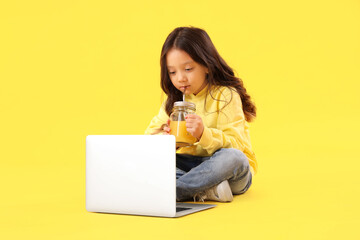 Canvas Print - Little Asian girl with mason jar of fresh citrus juice and laptop sitting on yellow background