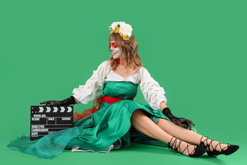 Poster - Young woman with painted skull on her face and clapperboard sitting against green background. Halloween celebration