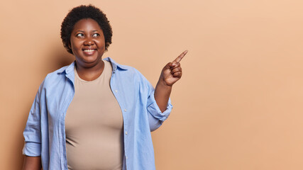 Overweight dark skinned woman with short curly hair points index finger aside shows place for your advertising text wears casual blue shirt isolated over brown background. People and promotion concept