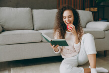 Young Smiling Happy Smart Woman Wear Casual Clothes Sits Near Grey Sofa Couch Read Book Novel Eat Apple Stay At Home Hotel Flat Rest Relax Spend Free Spare Time In Living Room Indoor. Lounge Concept.