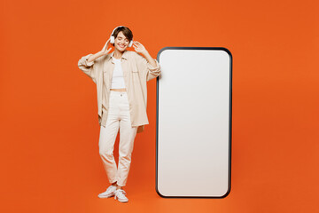 Wall Mural - Full body young happy woman wear beige shirt casual clothes big huge blank screen mobile cell phone smartphone with area listen to music in headphones isolated on plain orange red background studio.