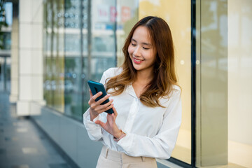 Wall Mural - A young woman is smiling while using her smartphone outside. She is holding the device with one hand and typing with the other. The woman is happy and connected.