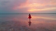 Aerial Of A Young Woman In Red Dress Walking In The Water Of A Unique Pink Salt Lake. Sunset At Lake Bursol With Beautiful Reflections On Calm Water Surface. Stunning Scenery