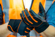 © Summer Paradive - Male construction worker with reflective orange vest putting black protective gloves on his hands. Workwear for builder, foreman or engineer