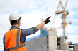 © Summer Paradive - Asian male construction worker or site engineer man with orange reflective vest and safety helmet pointing at unfinished building structure and tower crane