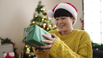 Poster - Young chinese woman holding gift standing by christmas tree at home