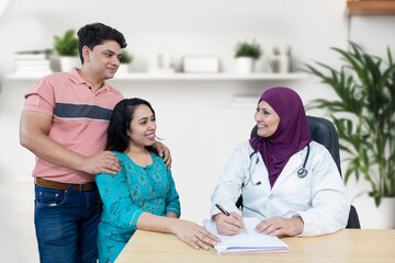 Wall Mural - Portrait of Indian female muslim doctor wearing head scarf consulting young married couple patients in fertility clinic about IVF or IUI. Planning pregnancy concept.