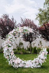 A beautiful wedding round arch, decorated with flowers, electric hanging lamps, stands on the green grass of a lawn in a garden, park. Festive photo.