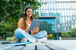 © Prostock-studio - Cool young woman student sitting outdoors, using digital tablet, headphones