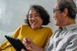 © Jirakul - Senior Asian couple sitting happily relaxing on the sofa, using tablet devices with hands, smiling and laughing together.