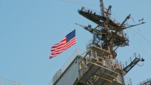American Flag Flies On The Mast Of A Warship. View Of Us Flag On Navy Ship. USA Flag And Antennas On Carrier Control Tower In Blue Sky. United States Warship Flying The American Flag