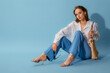 Fashionable young woman wearing  white linen shirt, trousers, trendy metallic strap sandals, carrying  wicker straw bag, posing on blue background. Studio portrait. Copy, empty space for text