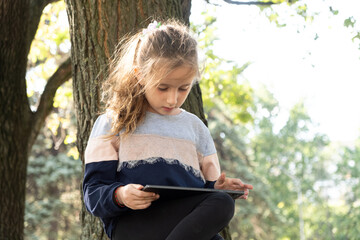 Wall Mural - Little girl uses a tablet while sitting in the park in the summer.