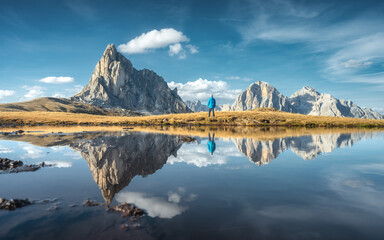 Wall Mural - Man and mountains reflected in lake at sunset in autumn. Passo Giau, Dolomites, Italy. Standing guy on the shore of lake is looking on high rocks and blue sky with clouds in fall. Reflection in water