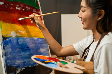 Wall Mural - Asian woman artist in the darkroom warm light working on painting with brush and variant acrylic color. Female artist painter on canvas in creative studio as art concept