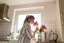 Woman Smelling Fresh Pink Flowers In Kitchen At Home
