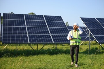 portrait of young indian man technician wearing white hard hat standing near solar panels against bl