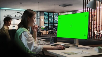 Fototapete - Creative Asian Woman Sitting at Her Desk Using Desktop Computer with Mock-up Green Screen Chromakey. Female Level Designer Working in Game Design Studio Diverse Office, Creating Immersive Gameplay.
