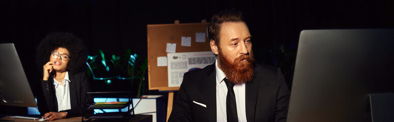 Wall Mural - bearded man working at night in office near tempting african american woman on background, banner