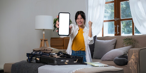 Wall Mural - Cheerful woman advertisement using mobile phone blank white screen display frameless modern design while pack clothes a suitcase ready for travel