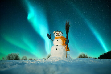 Wall Mural - Funny snowman in stylish red hat and red scalf on snowy field against the backdrop of incredible starry sky with Aurora borealis. Amazing night landscape. Northern lights in winter field