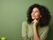 © Got Pink? - Headshot portrait of thoughtful pensive young brunette woman with curly hair holding hand under chin looking upward against honeydew-green studio wall background with copy space for text advertisement
