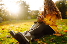 A Smiling Woman Sits Outdoors On The Lawn Among Yellow Autumn Leaves With A Phone In Her Hands. A Female Uses A Smartphone In An Autumn Park, Enjoying Nature.