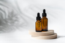 Cosmetic Serum Glass Bottles On Wooden Round Podium And White Background With The Shadow Of Plants. Organic Beauty Products Concept