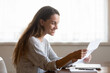 © fizkes - Smiling young woman reading pleasant news in letter, holding paper sheet, checking post mail, sitting at table at home, happy girl received refund, great shopping offer or exam results
