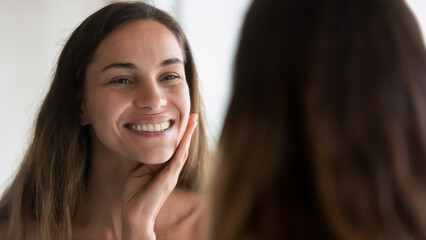 Wall Mural - Head shot smiling young woman with perfect healthy smooth skin touching cheek, looking in mirror, standing in bathroom after shower, doing face massage, skincare and natural beauty concept