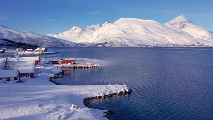 Wall Mural - Snow covered fishing village on coastline in winter, Norway. Surroundings of town Tromso. Panoramic aerial view landscape of nordic mountains, houses, rorbu, boats and ocean. Troms county, Fjordgard