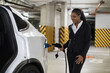 Beautiful young lady in formal attire attaching charger cable to white EV while standing in bottom-level garage. African american business person making use of parking time for getting auto filled up.