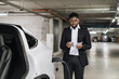 © sofiko14 - African male adult in business suit thumbing through cash while supplying battery-driven vehicle in indoor charge point. Handsome parking customer reminding of recent spending for EV charging.