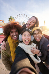 Vertical excited smiling group multiracial friends taking funny selfie looking at camera standing together outdoor. Multi-ethnic young people in amusement park having fun in sunny winter holiday. 