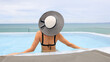 Back view of the young woman in a black bikini with a straw hat on the sun-tanned slim, shapely body with her arms spread to the side, relaxing in the swimming pool on the rooftop of the hotel, enjoyi