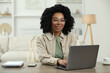 © New Africa - Happy young woman with earphones using laptop at wooden desk indoors