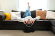 Happy young Asian woman relaxing at home, lying on the couch upside down, listening to music with headphones.