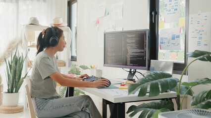 Professional young Asia lady IT development programmer listen music enjoy work type on keyboard coding programming fixing database on computer screen and laptop on table in workroom at house office.